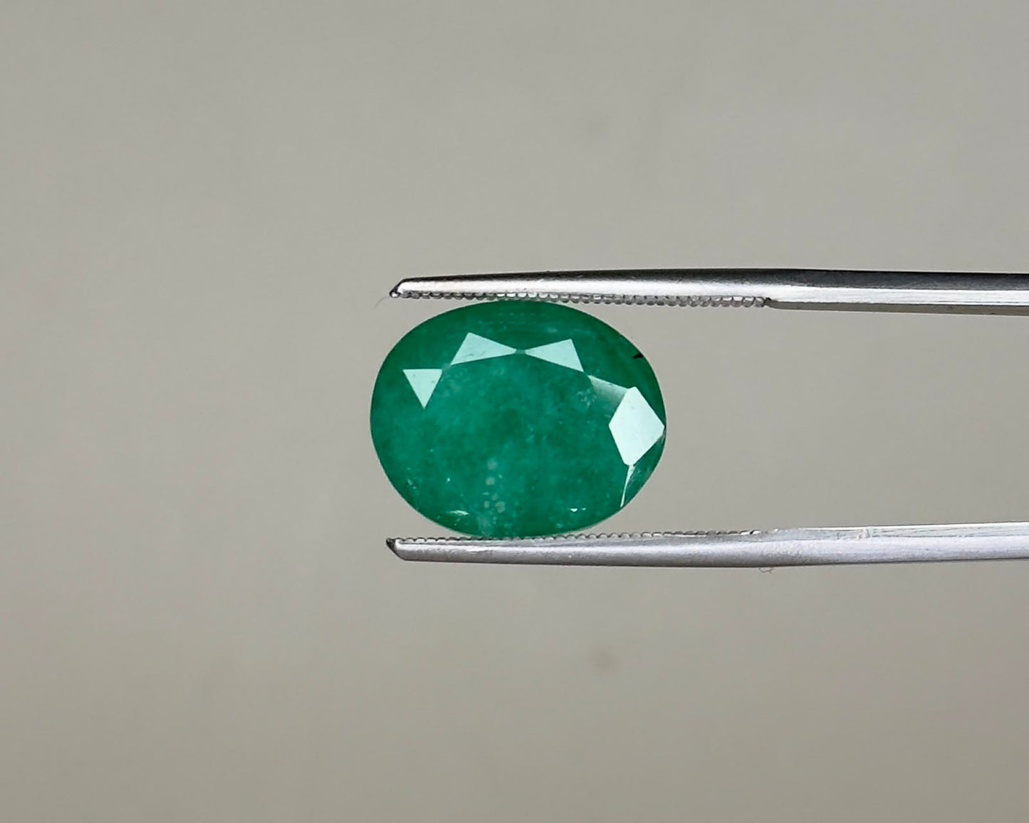 Emerald green oval gemstone with facets shown on metal tweezers