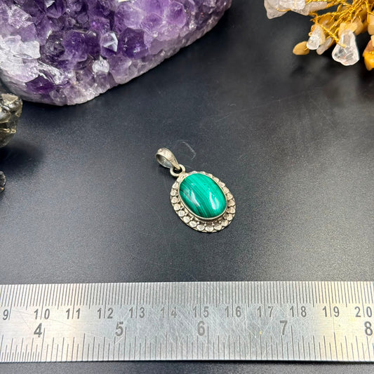 Malachite green oval pendant with textured silver border and bail on dark background with ruler