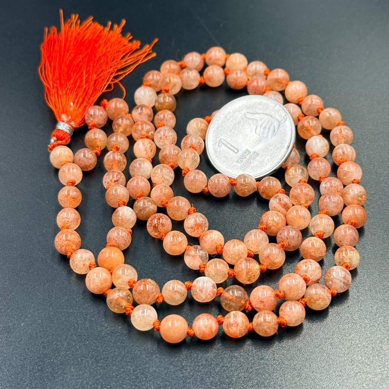 Close-up of sunstone mala 6mm orange bead bracelets with red tassel and coin for scale