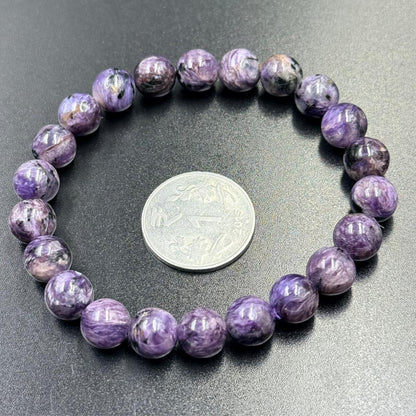 Charoite Bracelet with glossy round purple beads arranged around a silver coin on dark surface