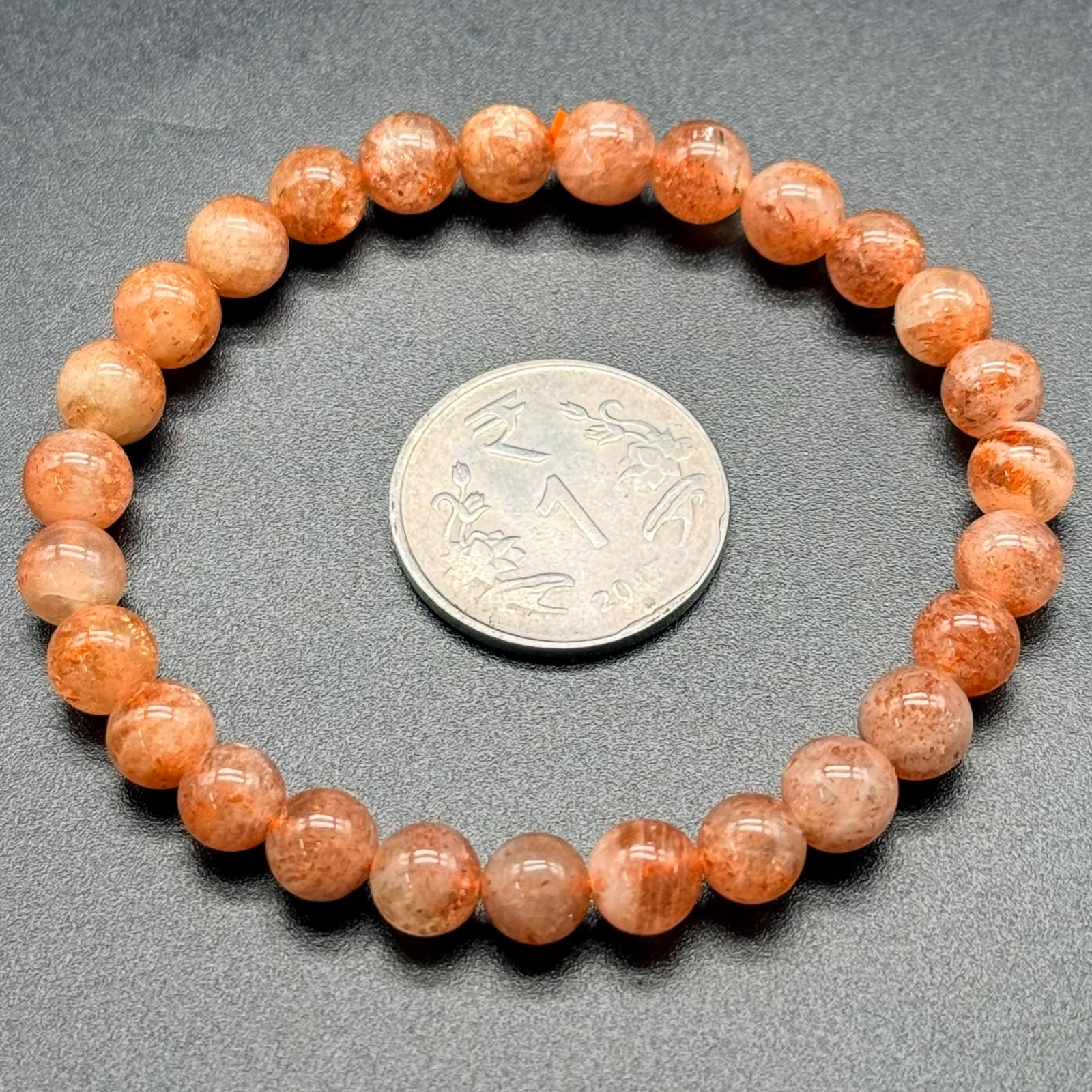 Sunstone Bracelet with round translucent orange beads arranged in a circle next to a one rupee coin on a gray surface
