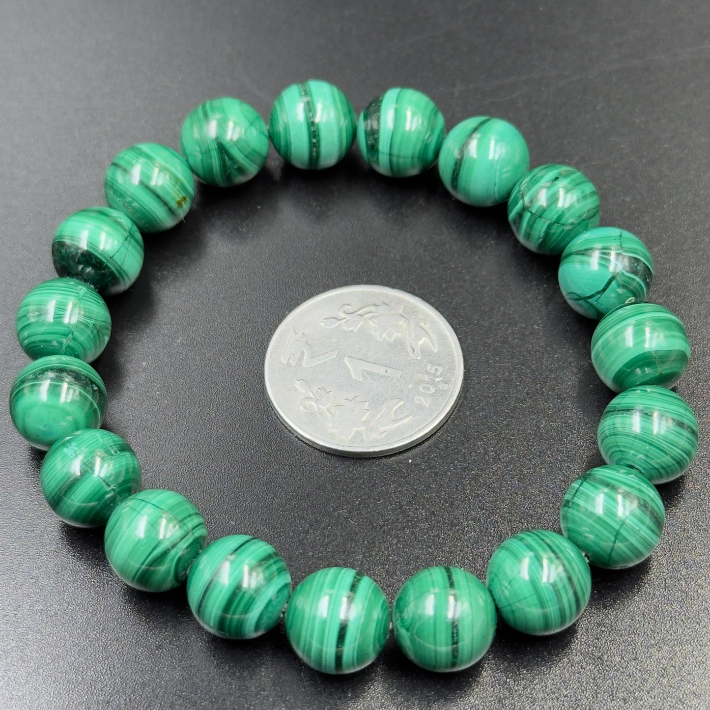 Malachite Bracelet with round green banded beads arranged around a silver coin on black surface