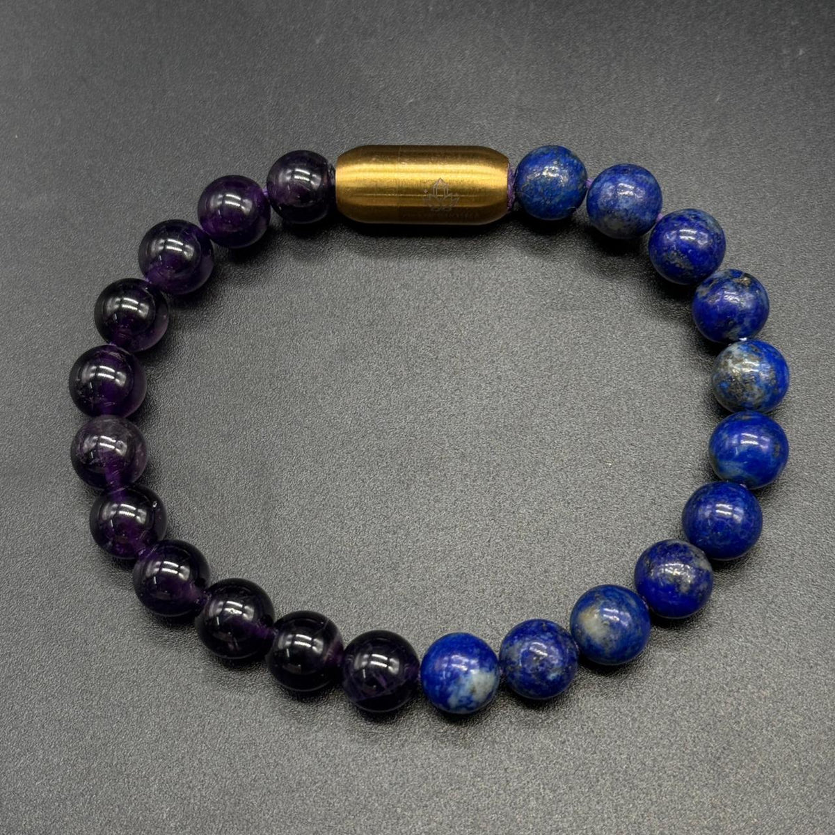 A beaded bracelet made of alternating dark purple amethyst and deep blue lapis lazuli ball beads, with a polished gold cylindrical accent bead at the top, displayed on a dark gray textured background  custom bracelet by GemKrishna