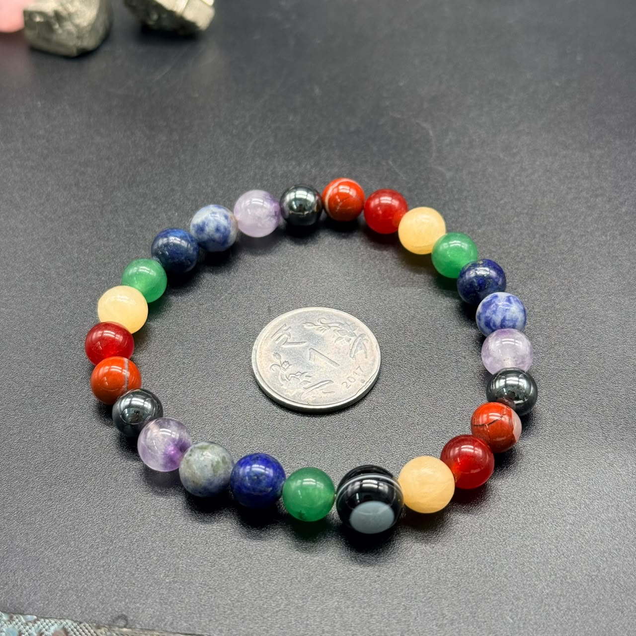 7 Chakra Balance Bracelet with multicolored round beads arranged in a circle next to a coin on a black surface