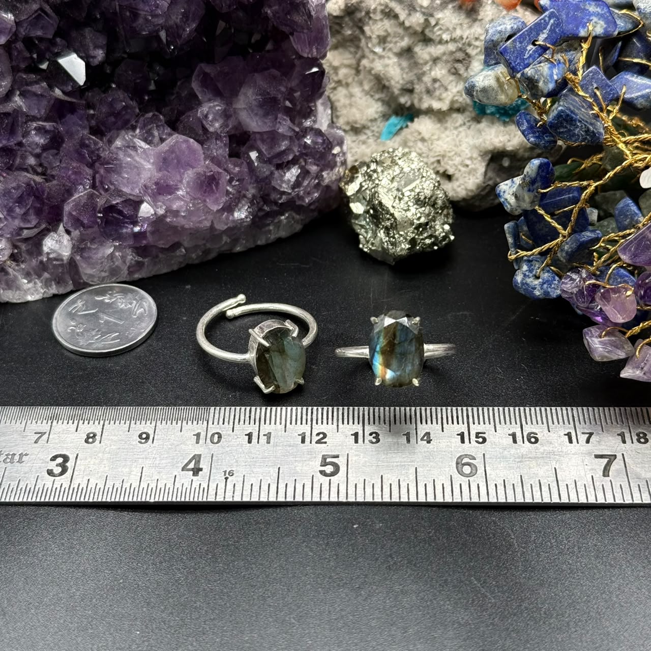 Two Natural Labradorite Rings with silver bands beside a metal ruler and gemstones on a black surface