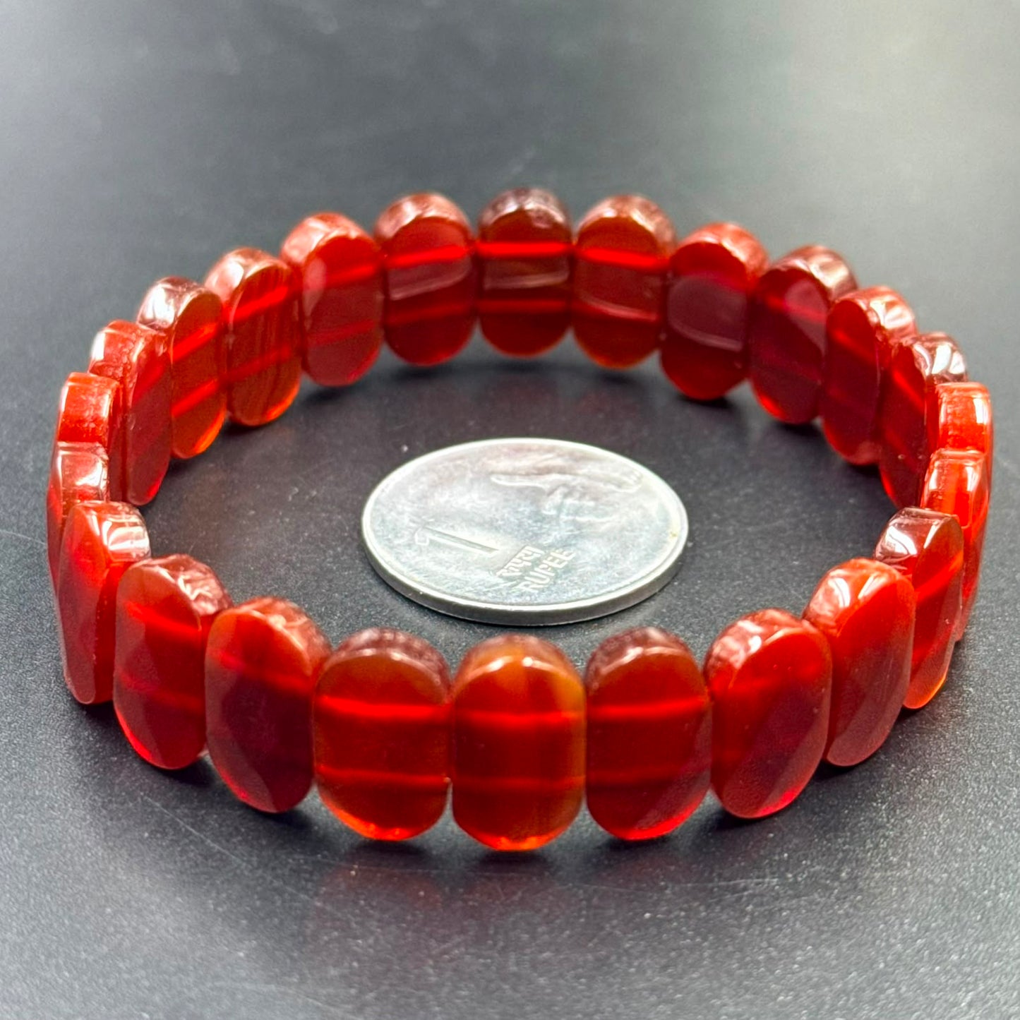 Carnelian flat oval bead bracelet displayed in a circle next to a 1 rupee coin on a dark surface