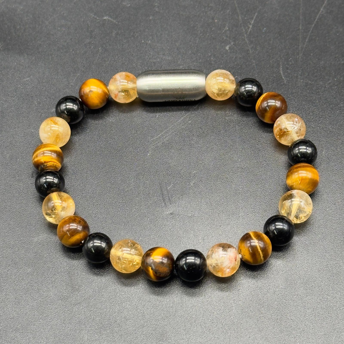 A beaded bracelet with alternating black tourmaline and tiger's eye beads, Citrine  crystals, and a central metallic silver lock magnetic , displayed on a dark gray textured surface.