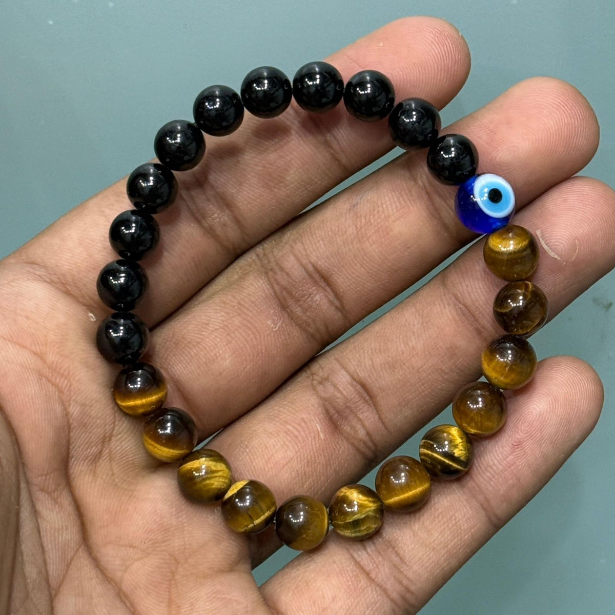 A beaded bracelet held in a hand, featuring alternating glossy black tourmaline and golden-brown tiger's eye beads, with a small blue evil eye charm near the top right.