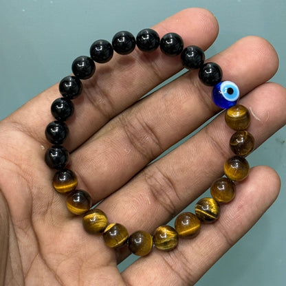 A beaded bracelet held in a hand, featuring alternating glossy black tourmaline and golden-brown tiger's eye beads, with a small blue evil eye charm near the top right.