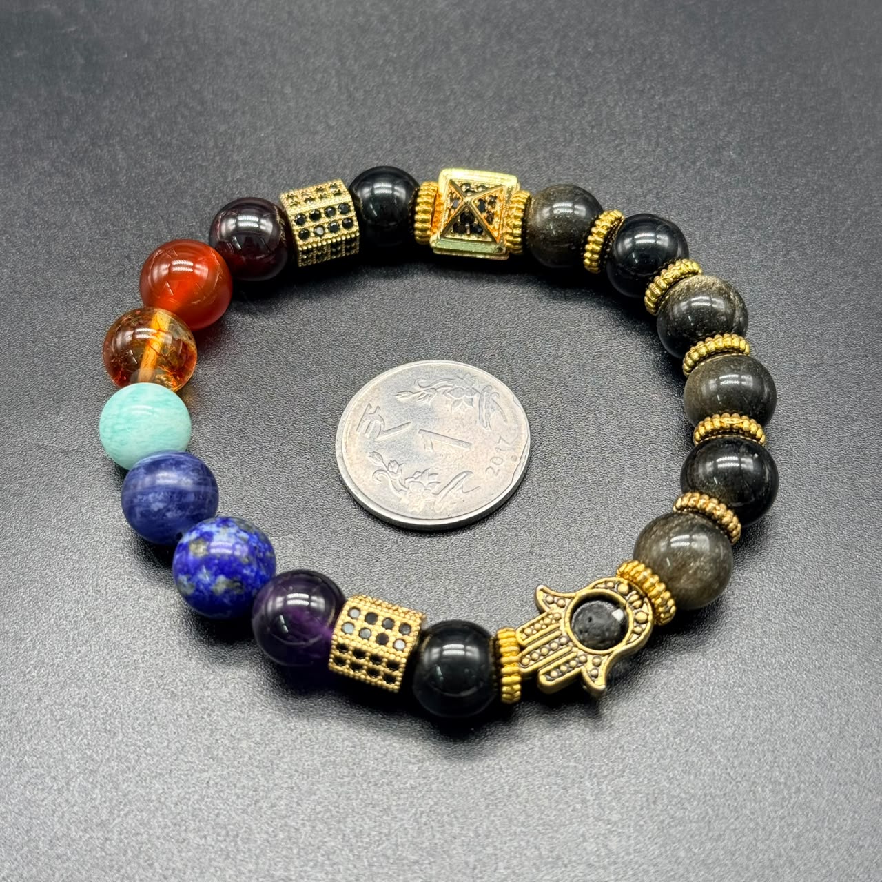 7 Chakra Balance Bracelet with multicolored beads, gold accents, and Hamsa hand charm next to coin