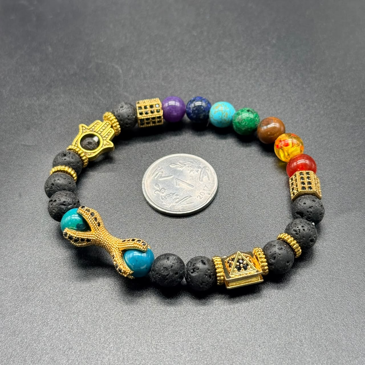 7 Chakra Balance Bracelet with multicolored beads, black lava stones, and gold accents next to a coin