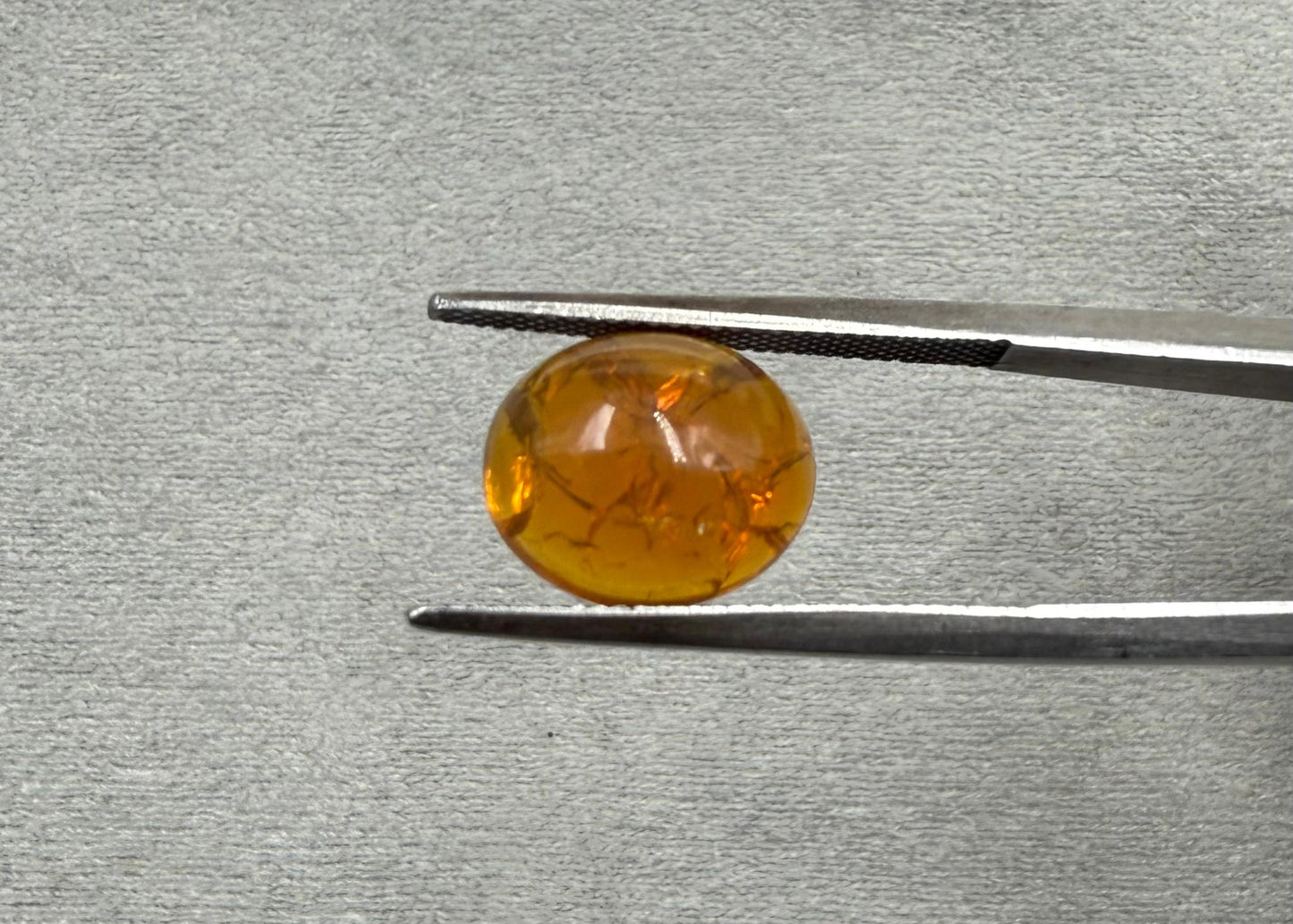 Round amber-colored gemstone held by tweezers on a gray textured background