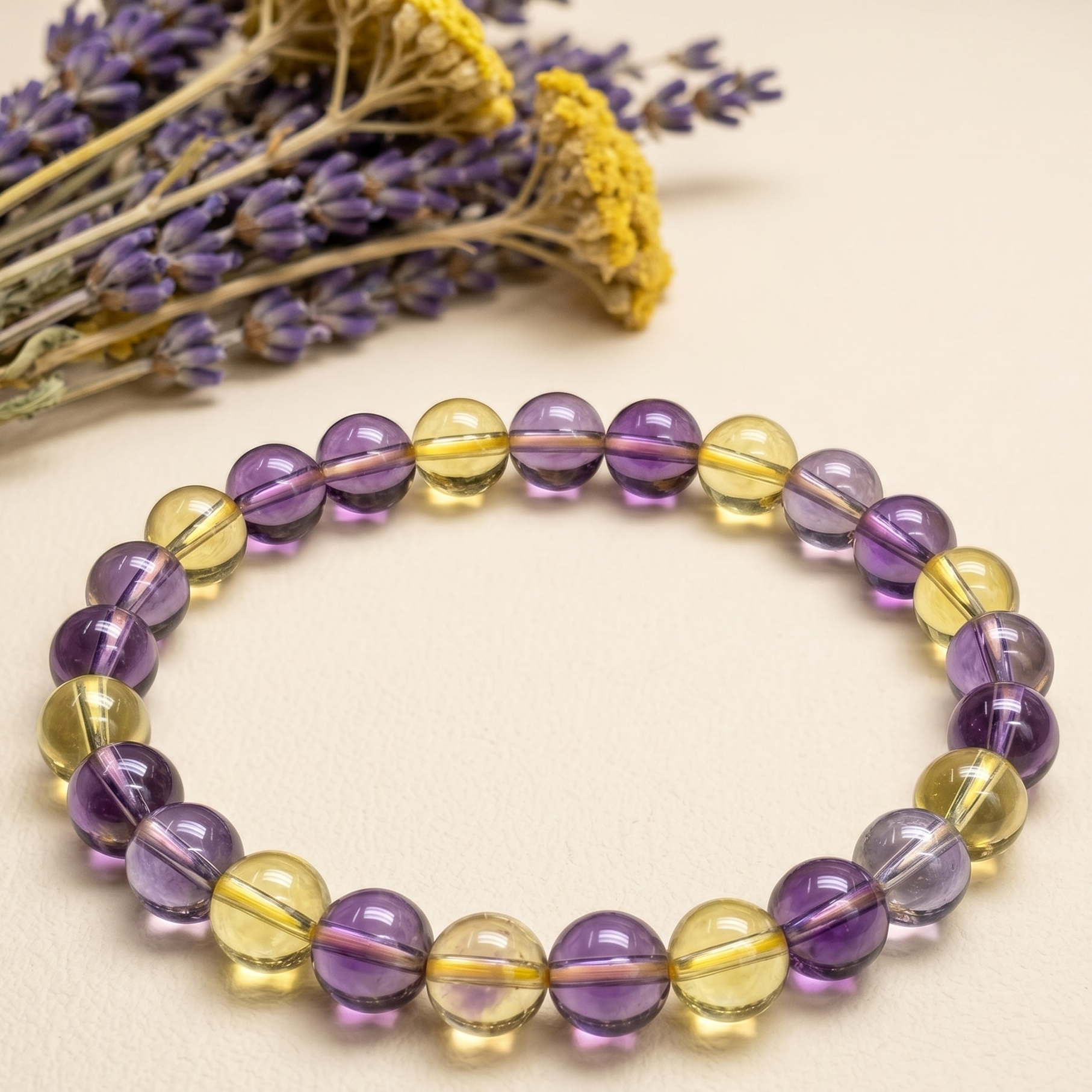 Ametrine Bracelet with alternating purple and yellow round beads on beige surface with dried flowers.