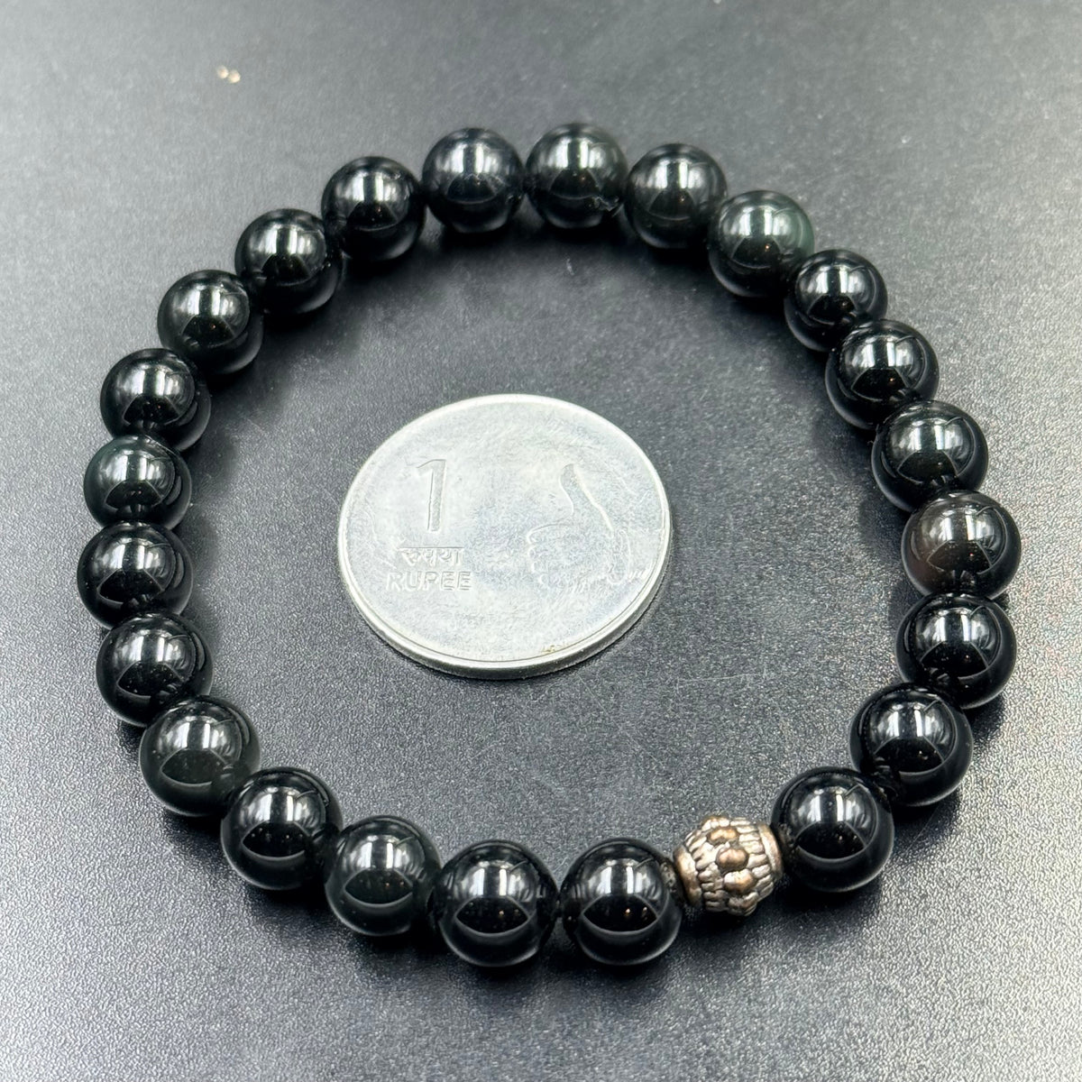 Close-up black obsidian bead bracelet with small silver-toned charm bead, with coin for scale