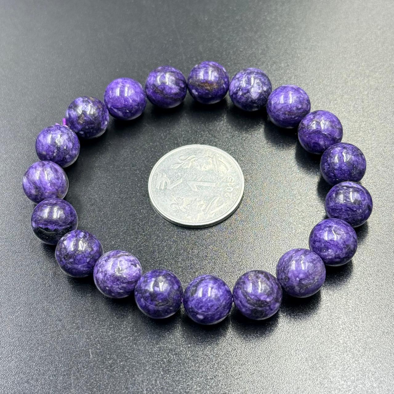 Charoite Bracelet with polished round purple beads arranged around a silver coin on black surface