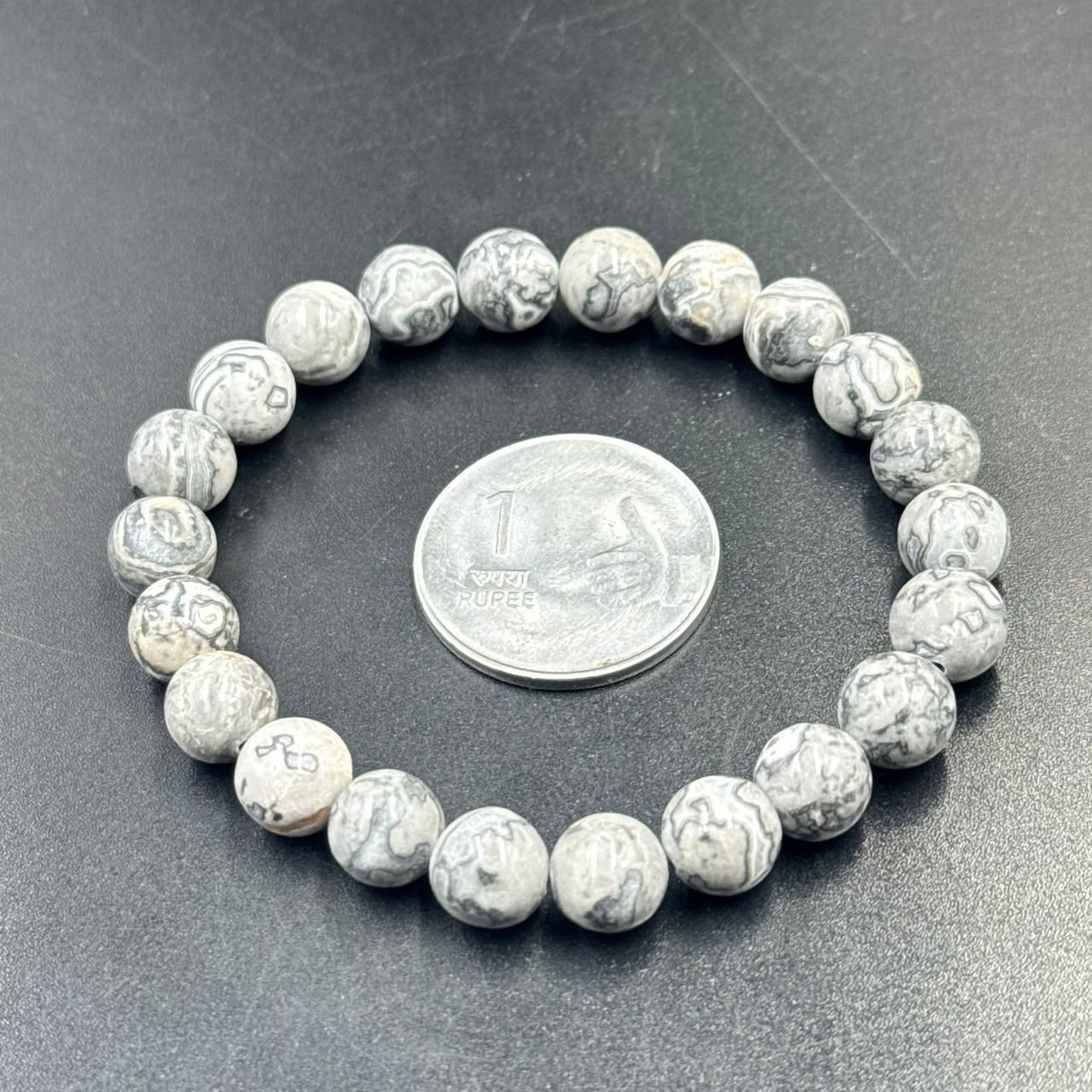 Mapstone Bracelet with round marbled gray and white beads beside a 1 rupee coin on black surface