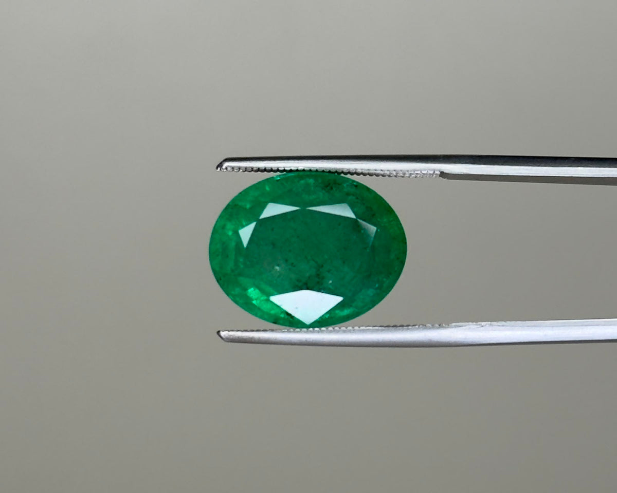 Emerald green oval gemstone with visible facets, held between metal tweezers