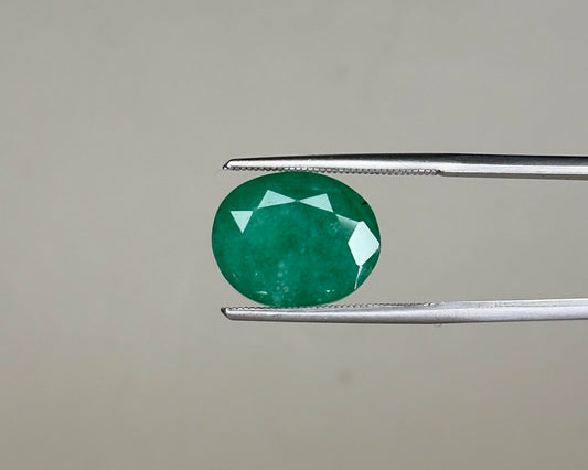 Emerald green oval gemstone with facets shown on metal tweezers