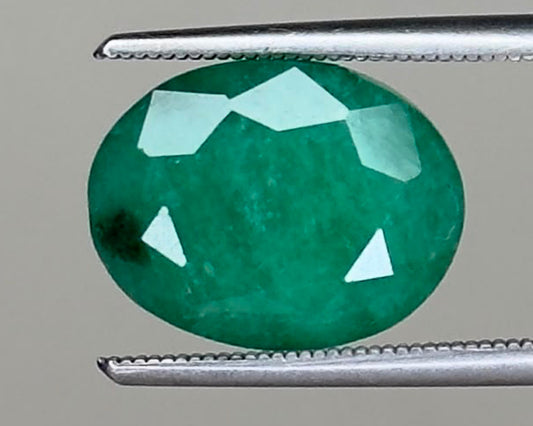 Emerald oval gemstone, 6.07 carats, held between metal tweezers on a gray background