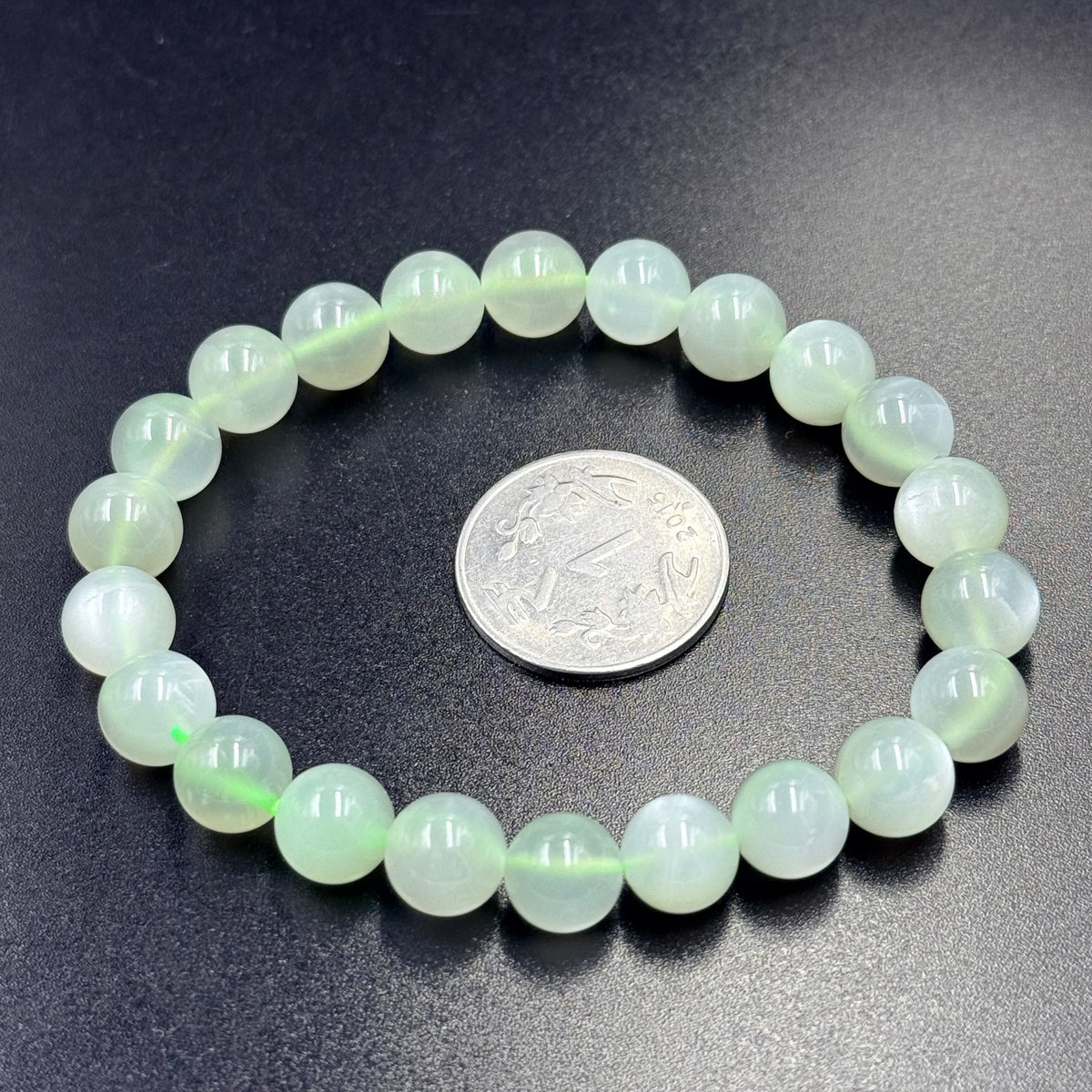 Green moonstone bead bracelet with round beads shown beside a silver coin on dark fabric