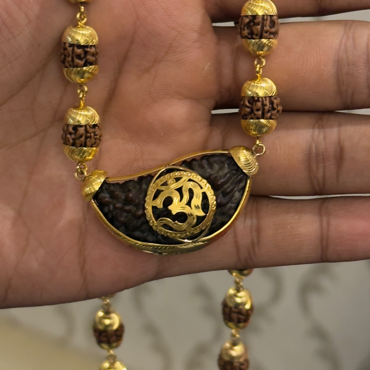 Close-up of hands holding a gold-plated rudraksha mala with Om pendant teardrop