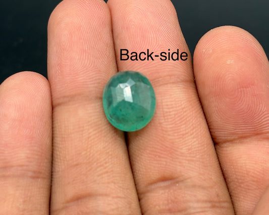 Natural emerald oval gemstone back-side view held between fingers, green faceted cabochon