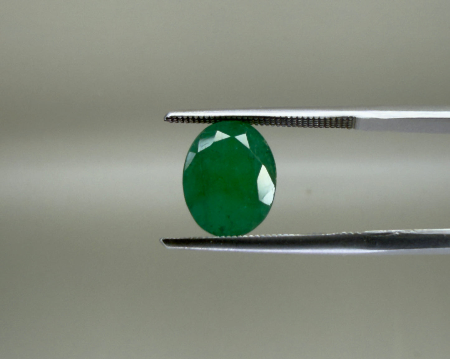 Natural emerald oval mixed cut gemstone held with tweezers on a gray background