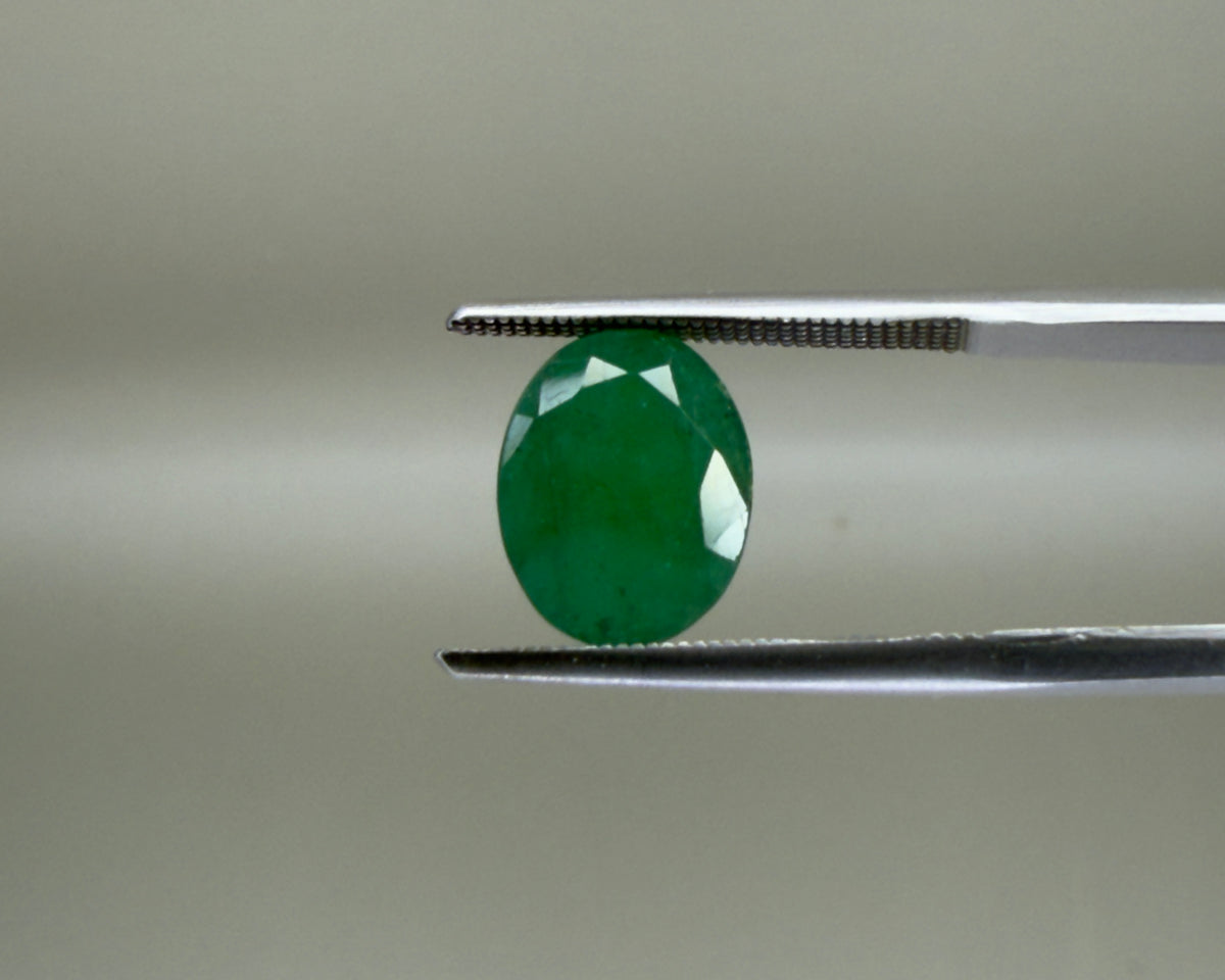 Natural emerald oval mixed cut gemstone held with tweezers on a gray background