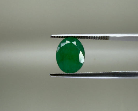 Natural emerald oval mixed cut gemstone held with tweezers on a gray background
