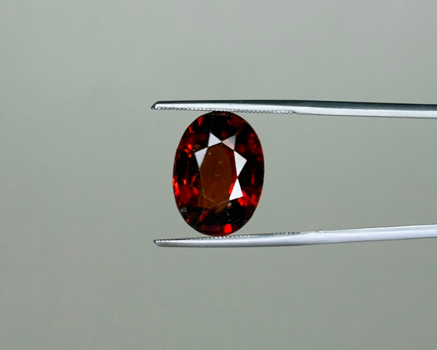 Natural hessonite garnet oval gemstone with red and dark facets held on tweezers