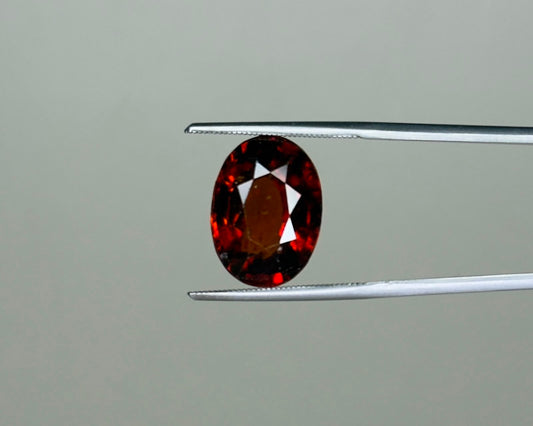 Natural hessonite garnet oval gemstone with red and dark facets held on tweezers