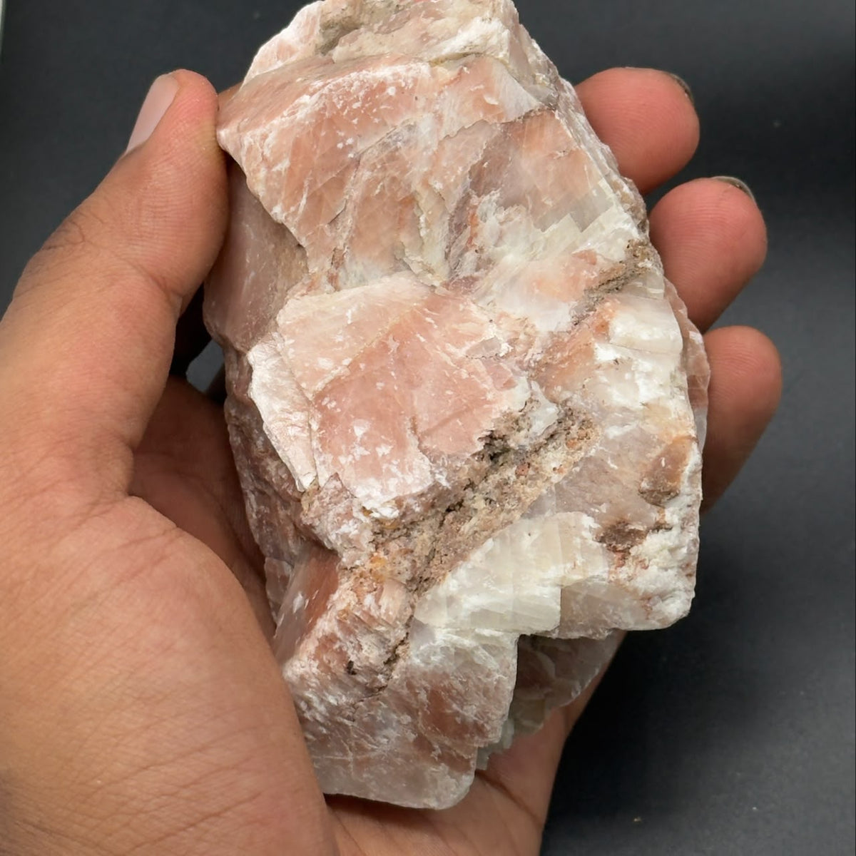 Natural sunstone rough crystal held in hand, showing peach and white facets on gray surface