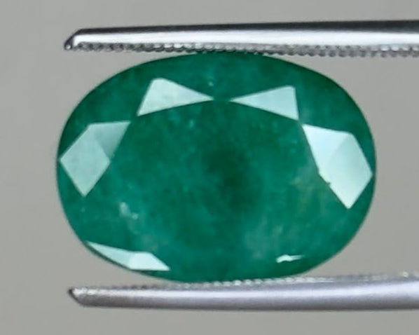 Emerald gemstone oval cut with bright green color held between tweezers