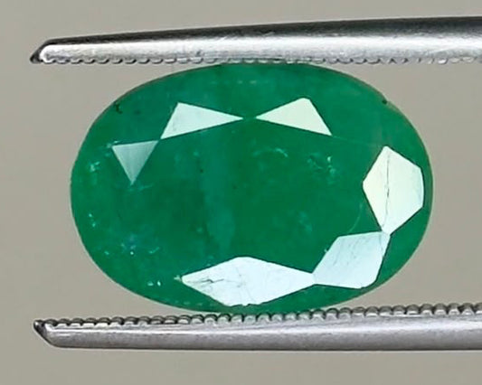 Emerald oval gemstone with visible facets, held between metal tweezers
