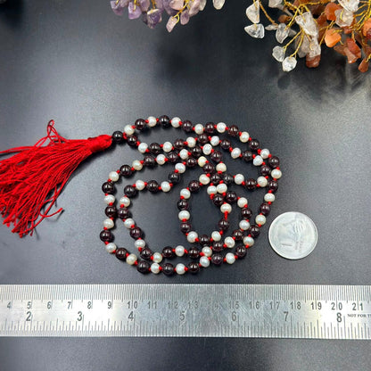 Pearl and garnet jaap mala beads with red tassel, laid out on a grey background with coin