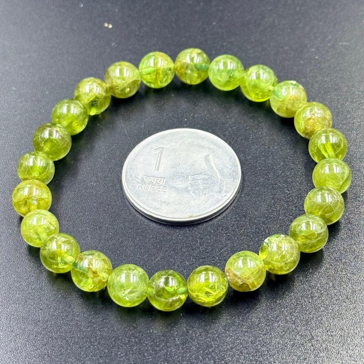 Peridot Gem Quality Bracelet with round green beads beside a one rupee coin on black surface