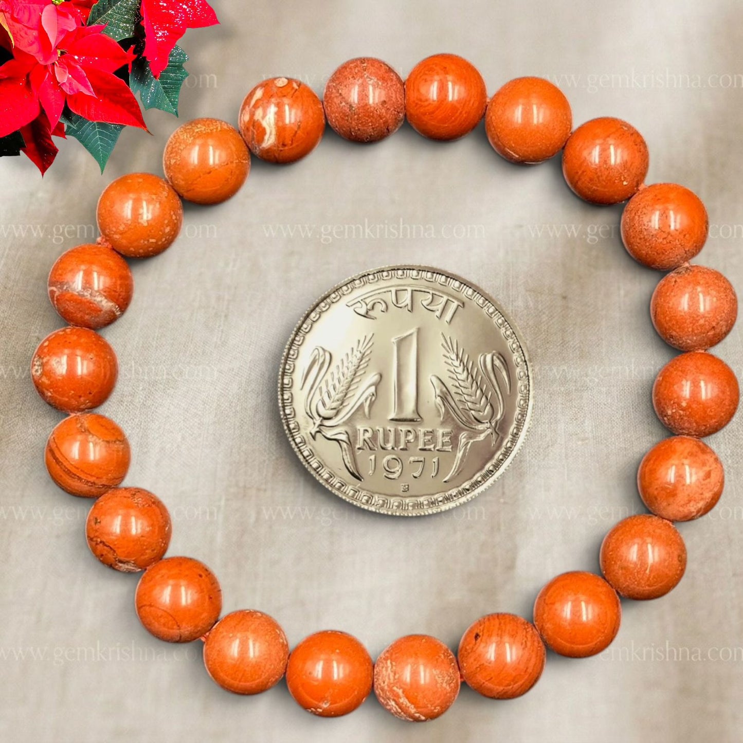 Red jasper bracelet beads with Rupee coin centerpiece on gray fabric and red flowers at top left