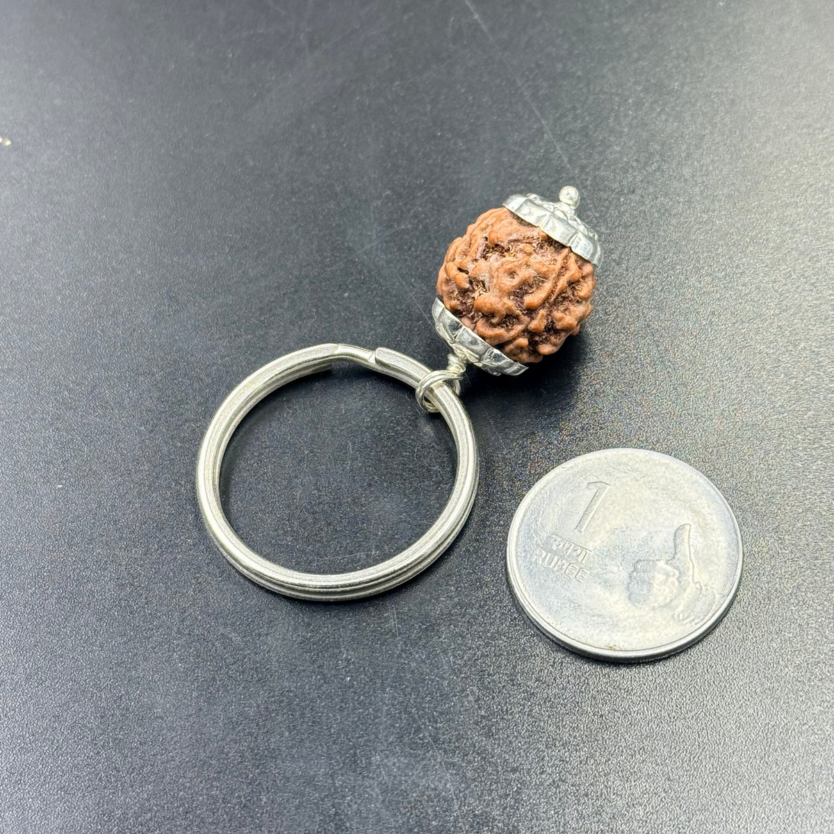 Rudraksha keychain pendant with silver-tone cap and metal ring next to coin on blue background