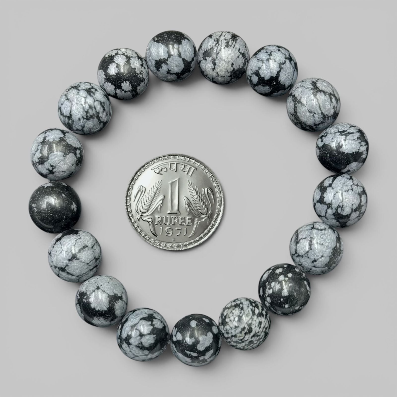 Snowflake obsidian bracelet with round black and white speckled beads beside a coin on gray background